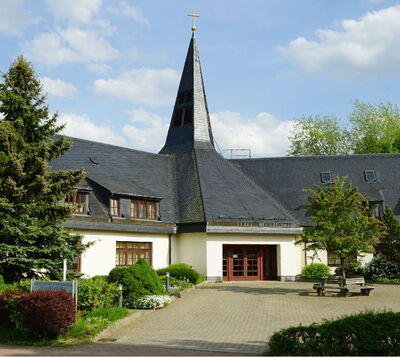 Kirche mit schiefergedecktem Spitzturm, Kreuz und gepflegtem Vorplatz mit Bepflanzung und Sitzbänken