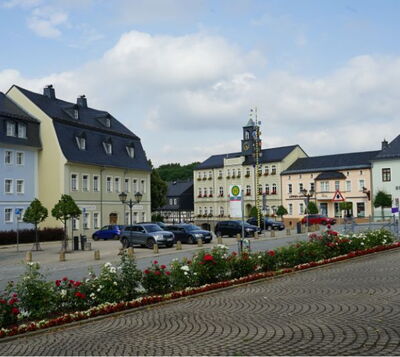 Historischer Marktplatz mit Rathaus, gepflasterter Fläche, Blumenbeeten und parkenden Autos vor pastellfarbenen Gebäuden.