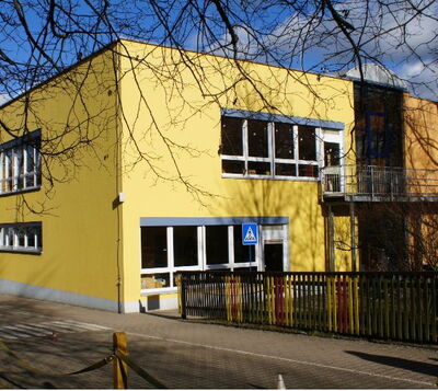 Moderner Zweckbau mit gelber Fassade, großen Fensterfronten und buntem Zaun im Eingangsbereich einer Schule